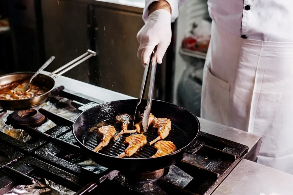 Cozinheiro Usando Pinça Para Grelhar Costeletas Em Fogão Industrial, Com Panela Ao Fundo.